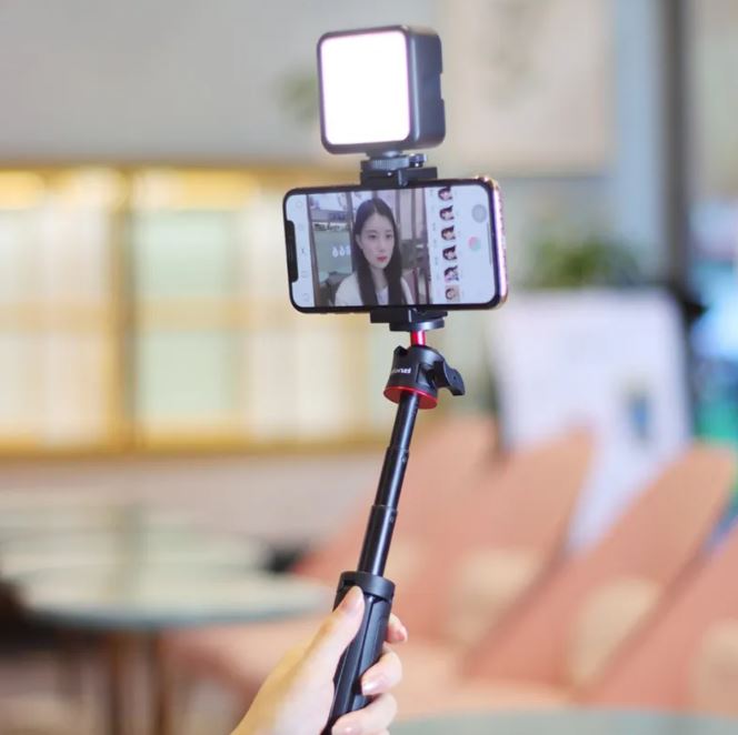 Black selfie stick with LED fill light mounted to a phone, held in hand