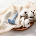 Light blue safety razor on a cloth with cotton flowers and soap nearby.